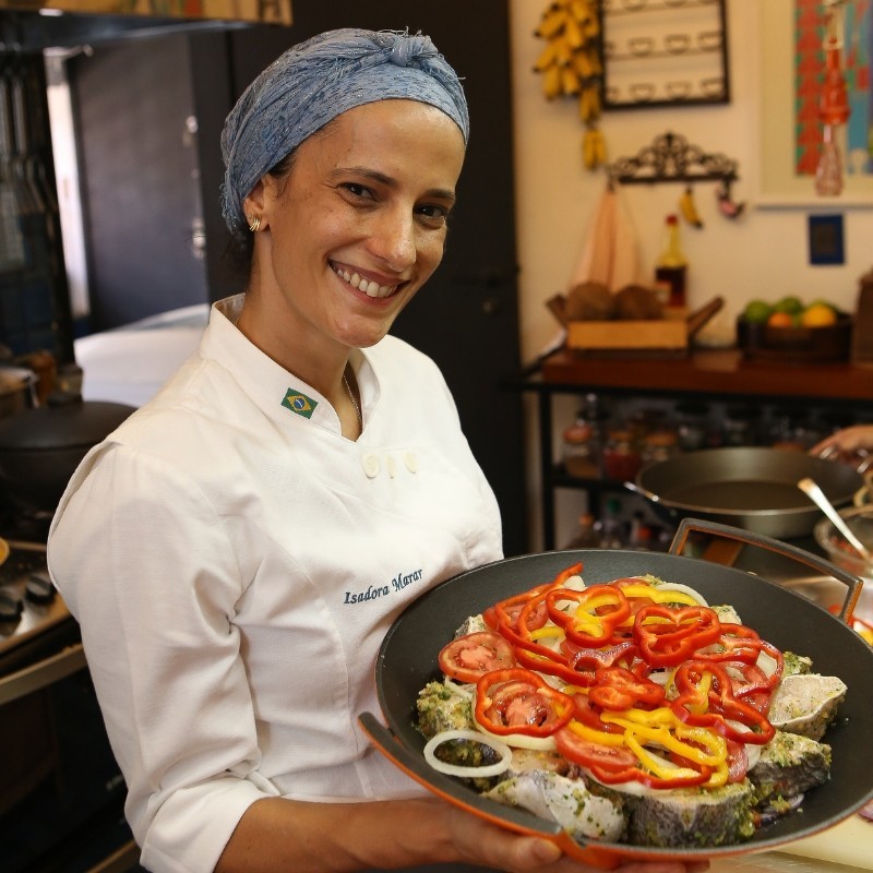 Chef Isadora Marar sorrindo com uma travessa de peixe temperado em sua cozinha